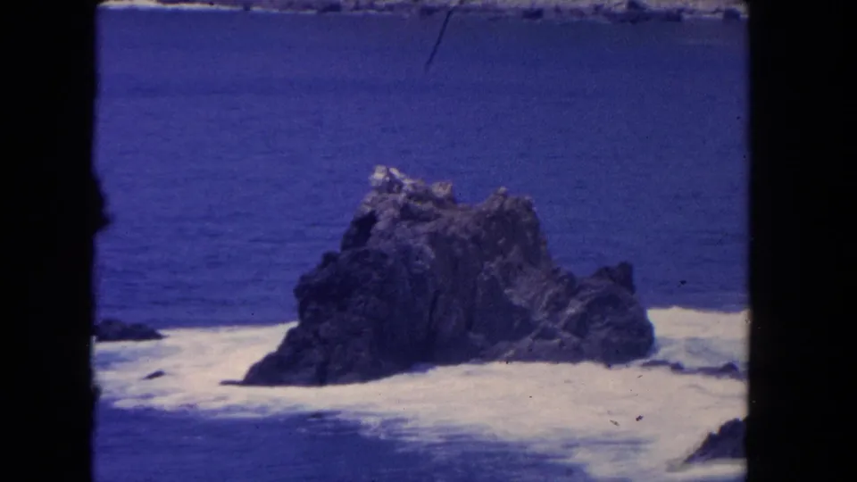 1962 Los Angeles coastline frame showing a rugged offshore rock with blue water and white surf around it.