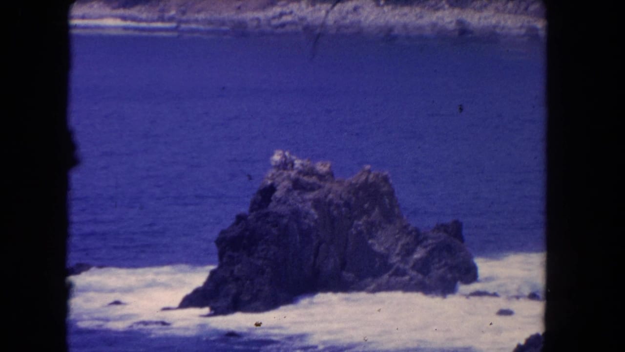 Dark rock formation in ocean surf under a blue sky.