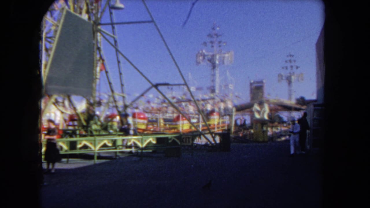Outdoor carnival midway with rides, metal supports, visitors, and a clear blue sky.