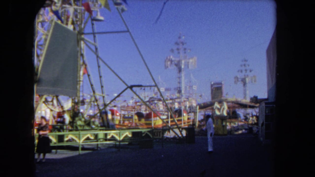 Carnival rides and midway booths with a woman in dark clothing and a person in white nearby.