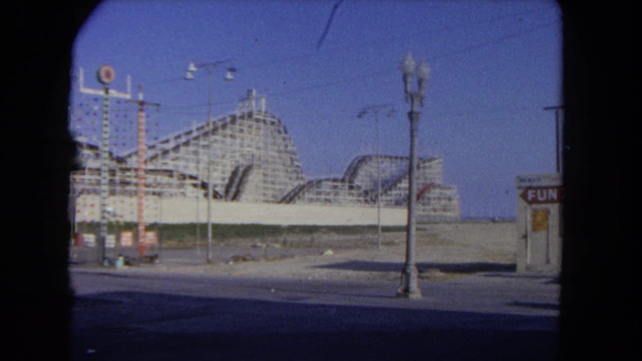 A large wooden roller coaster rises beyond lamp posts and open midway space in 1962 Los Angeles amusement park footage.