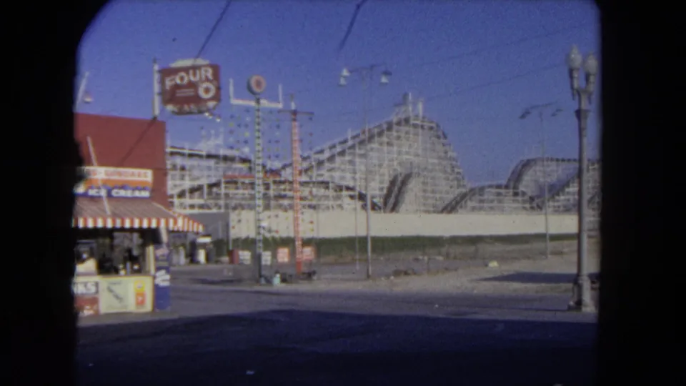 1962 Los Angeles amusement-park view with a red ice cream stand, striped awning, roadside signs, and wooden roller coaster behind it.