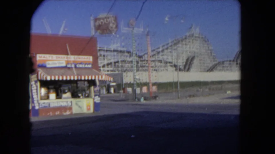 1962 Los Angeles 8mm frame showing a concession stand with business signs in front of a large wooden roller coaster.