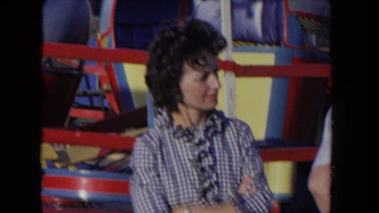 Woman in a checkered blouse stands with arms crossed in front of a colorful carnival ride.