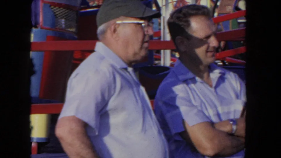 Two men, one wearing a cap and glasses, stand near the colorful ride structure in 1962 Los Angeles home-movie footage.