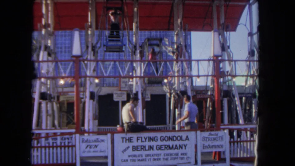 At a 1962 Los Angeles fair, two men stand by The Flying Gondola ride entrance as gondola seats hang above the signed platform.