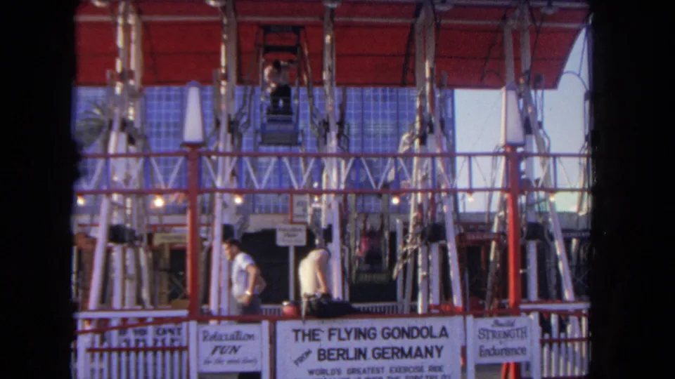 A visitor passes through the foreground while another man remains near The Flying Gondola sign and the ride machinery above.