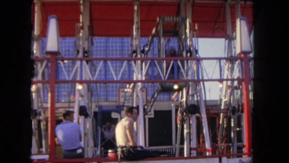 The home-movie camera frames two adult men beside The Flying Gondola ride, with the red canopy and metal supports above.