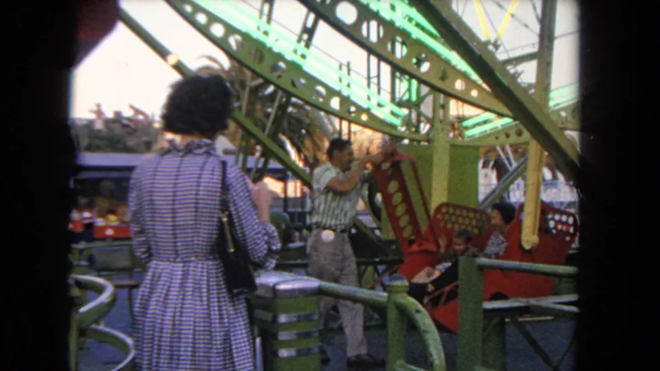 Home-movie frame of a fairground attendant helping at a red Ferris wheel gondola as a woman stands nearby in 1962 Los Angeles.