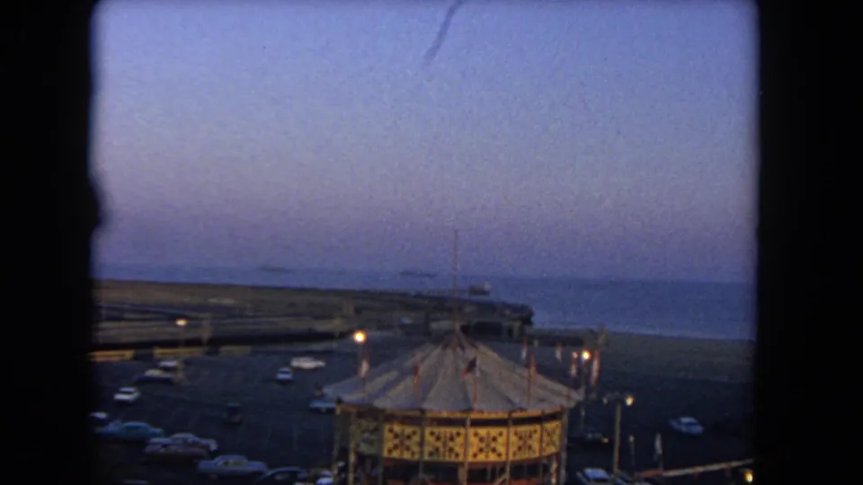 1962 Los Angeles home-movie view over an oceanside carnival, with a carousel roof, cars, and ocean beyond at dusk.