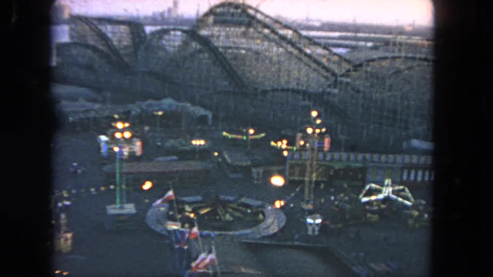 1962 Los Angeles 8mm frame showing an amusement park at dusk, including a roller coaster, flags, and glowing ride areas.
