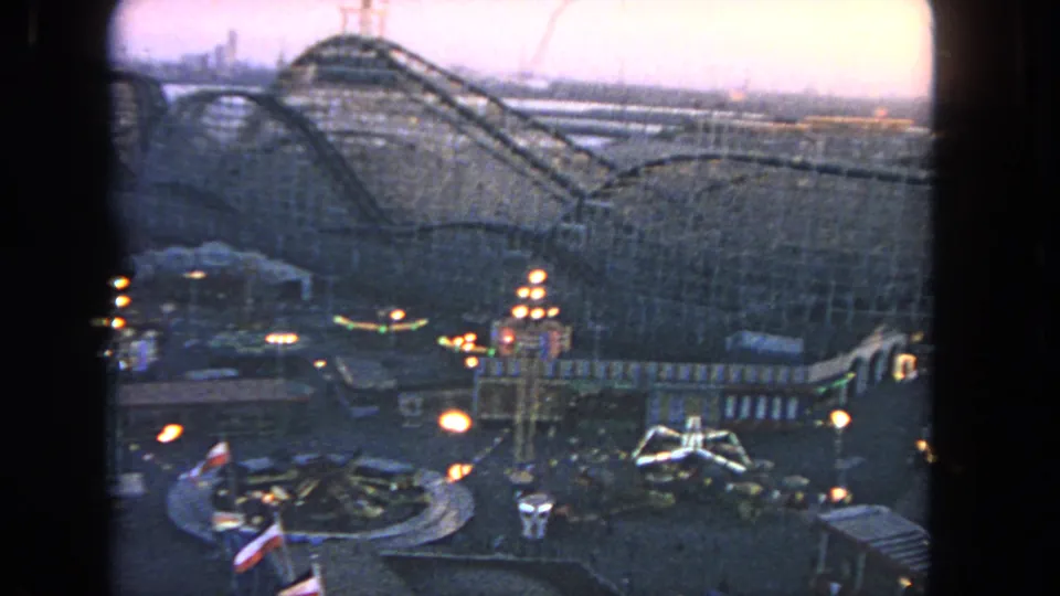 End frame of a 1962 Los Angeles amusement-park view, with wooden coaster tracks, lighted attractions, and evening sky.