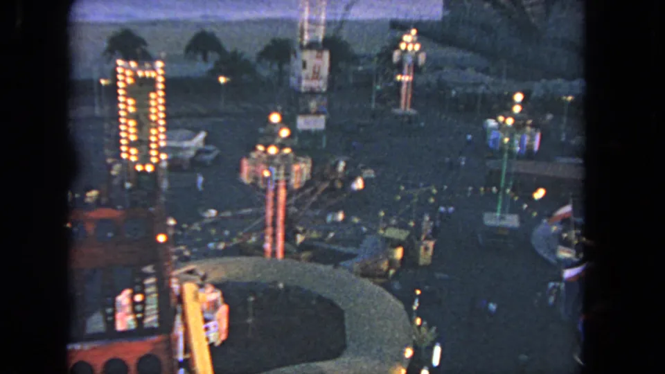 1962 Los Angeles 8mm frame showing illuminated midway rides, palm trees, walkways, and distant water from above.