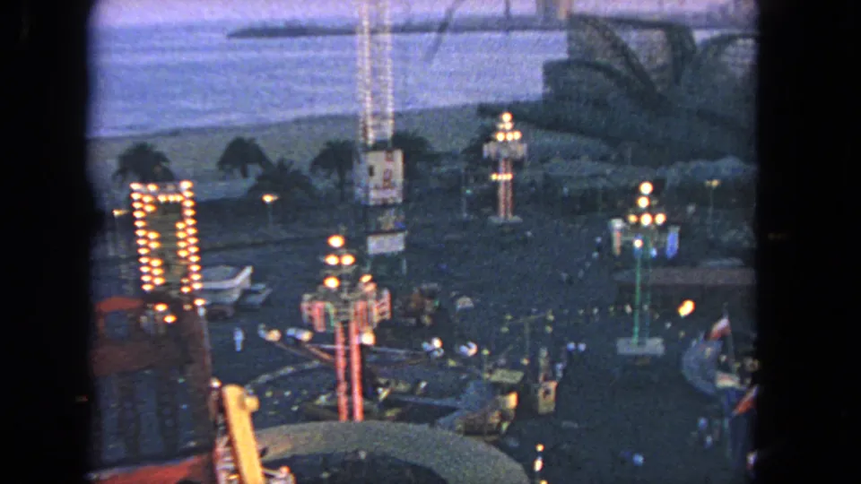 High 1962 Los Angeles amusement-park view with ride lights, small visitors, palm trees, and water beyond the midway.