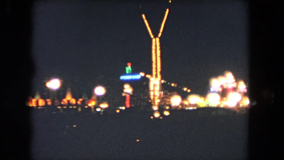 Nighttime Los Angeles fair or amusement scene with a tall lit tower, glowing lamps, and soft distant attractions against a dark sky.