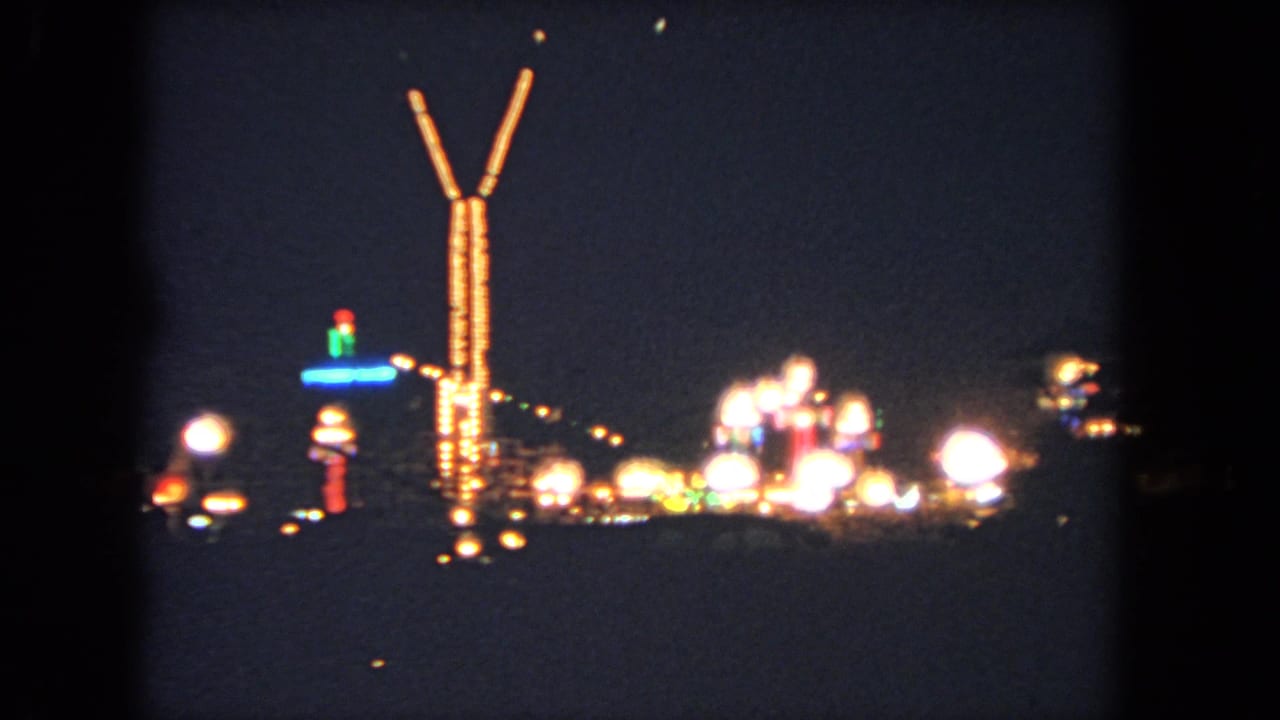 Distant nighttime amusement lights with a tall illuminated V-shaped tower against a dark sky.