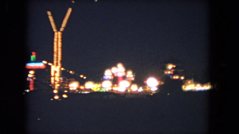1962 Los Angeles home-movie frame with blurred amusement lights, a V-shaped tower, and bright circular bulbs in the distance.