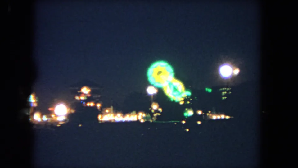 1962 Los Angeles 8mm frame of distant fairground lighting, with a green and yellow illuminated ride near the right side.