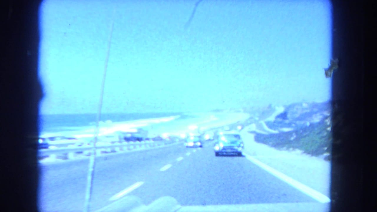 1962 Los Angeles coastal driving view from inside a car, showing traffic, ocean, guardrail, and blue-shifted 8mm color.