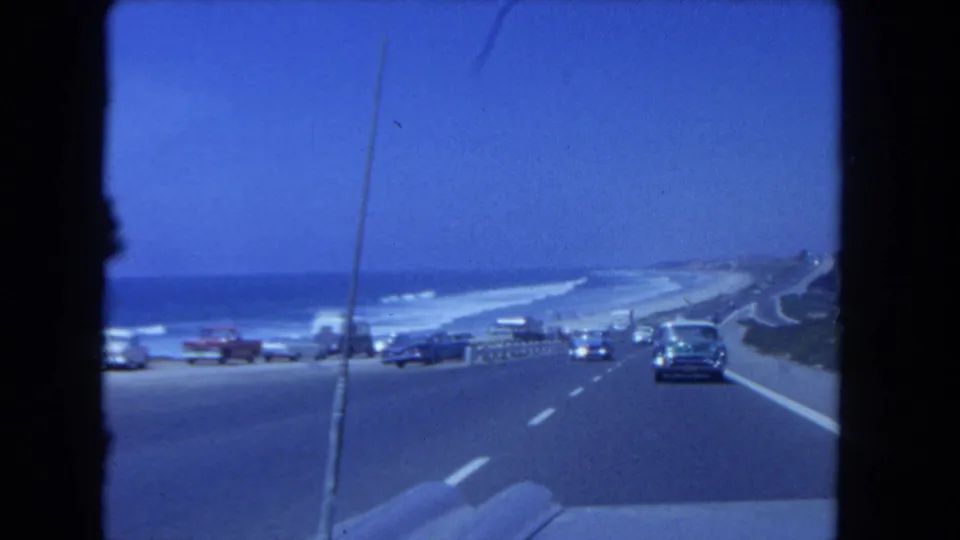 Mid-century cars travel along a Los Angeles coastal road beside the beach and ocean, filmed from a moving vehicle on 8mm.