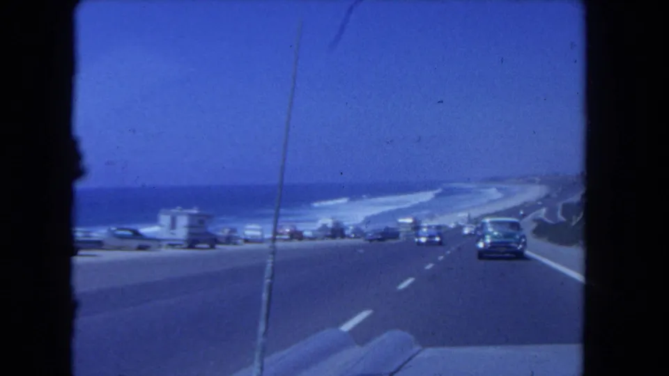Los Angeles coastal highway traffic passes beach parking and a trailer near the ocean in a blue-toned 1962 8mm home-movie frame.