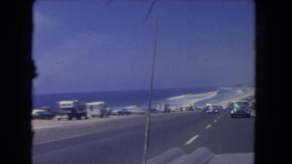 1962 Los Angeles-area coastal road scene with parked vehicles, camper trailers, traffic, ocean, and blue sky from a moving car.