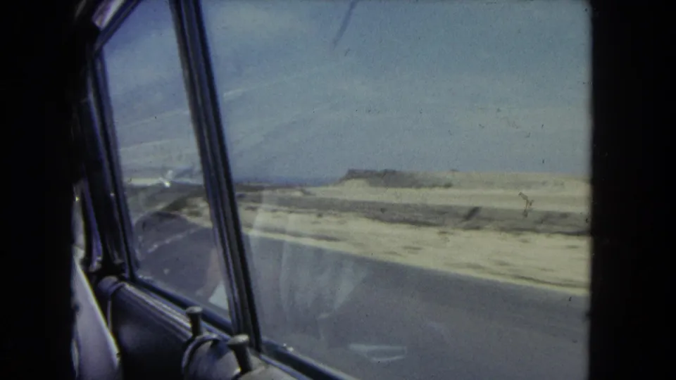 Passenger-window view during a 1962 Los Angeles coastal drive, with sandy roadside terrain, distant low structures, and car interior framing.