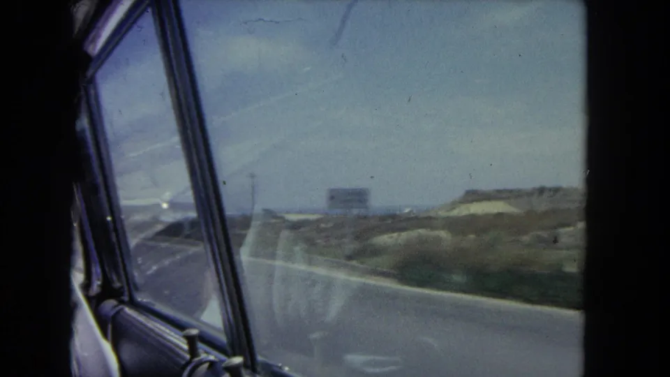 1962 Los Angeles-area 8mm passenger-window view of a coastal road bordered by sandy dunes and sparse vegetation.
