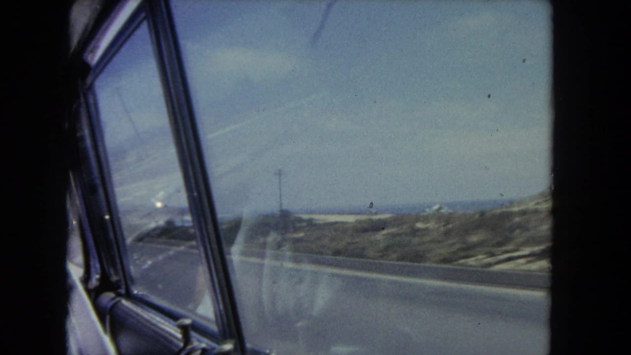 Passenger-side view from a moving car along a 1962 Los Angeles coastal road, with dunes and ocean horizon visible.