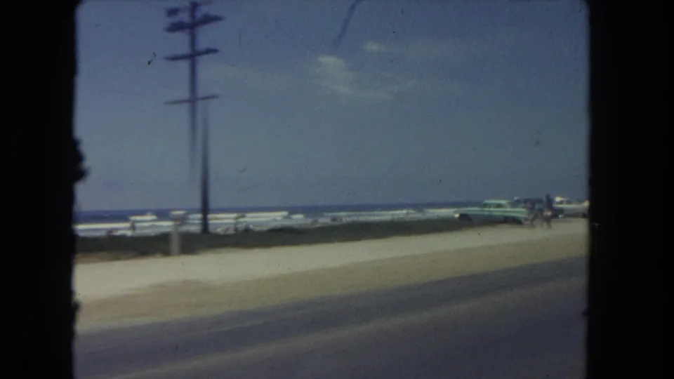Coastal Los Angeles road scene from 1962 showing beach parking, a utility pole, ocean horizon, and distant people.