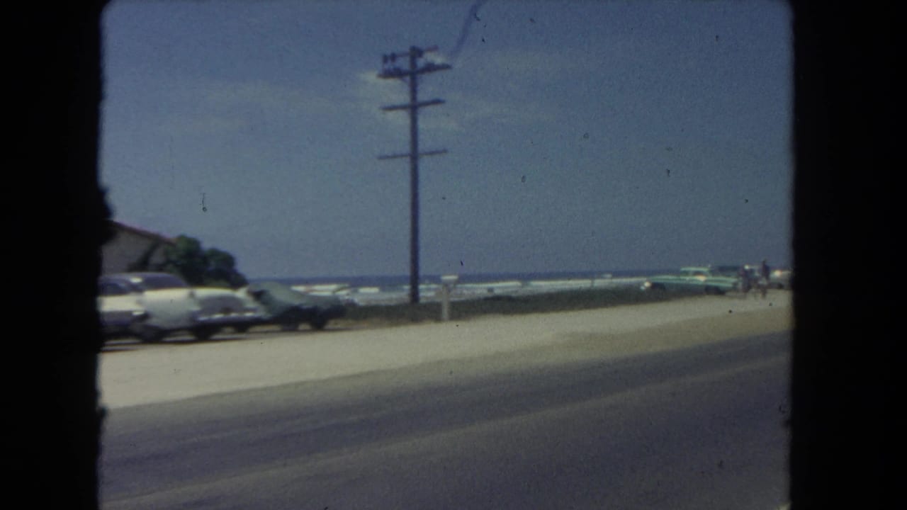 1962 Los Angeles coastal roadside view from a moving vehicle, with parked cars, a utility pole, beach sand, and ocean surf beyond.