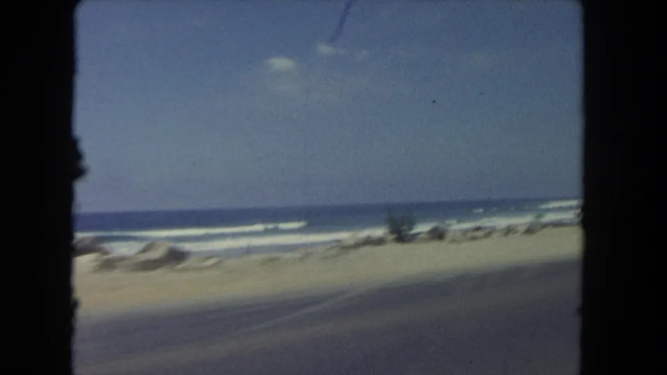 1962 Los Angeles beachside road view with ocean surf, sand, rocks, sparse plants, and the roadway blurred by vehicle motion.