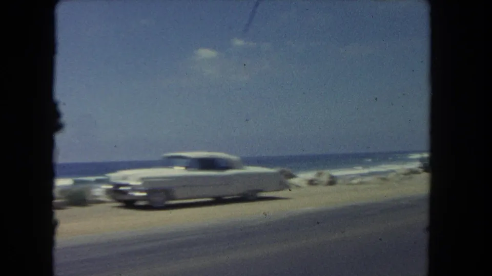 A white early-1960s sedan passes along a Los Angeles-area coastal road, with blue ocean, surf, shoreline rocks, and dark frame edges visible from a moving vehicle.