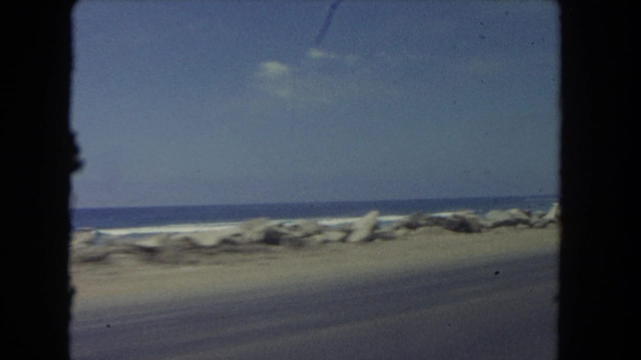 Empty stretch of coastal road with rocks, surf, ocean, and blue sky.