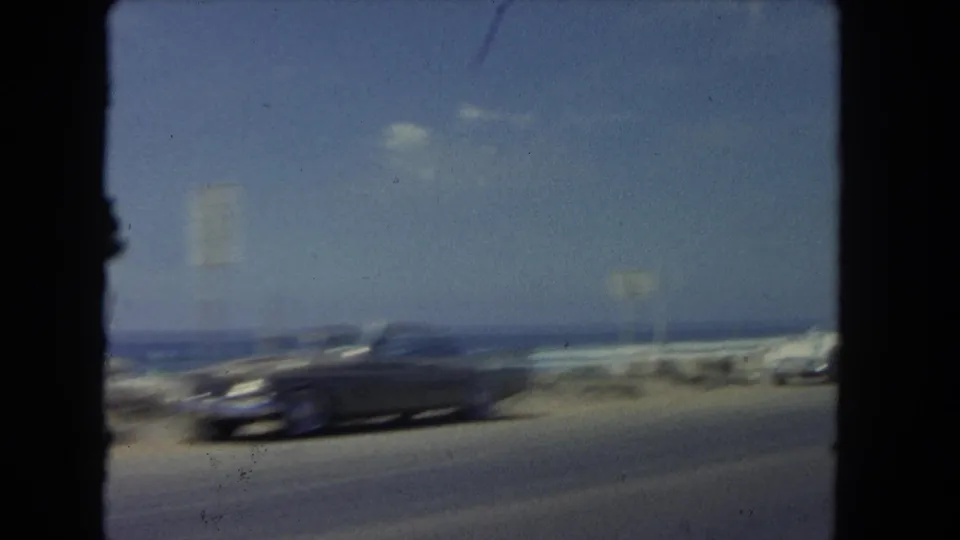 A dark mid-century sedan is motion-blurred as it passes along a Los Angeles-area coastal road, with ocean, rocks, surf, and blue sky in the background.