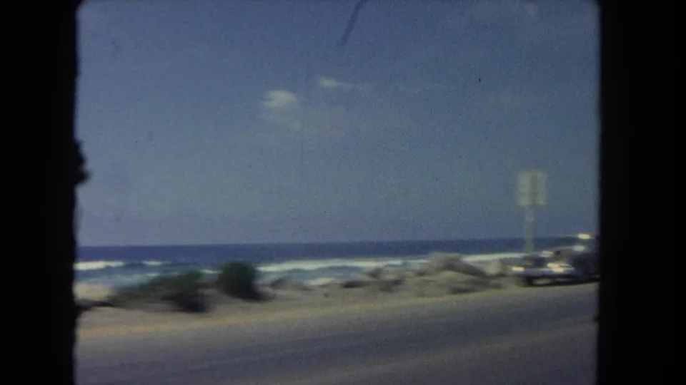1962 Los Angeles coastal road view from a moving car, with ocean surf, roadside posts, and a blurred dark vehicle near the shore.