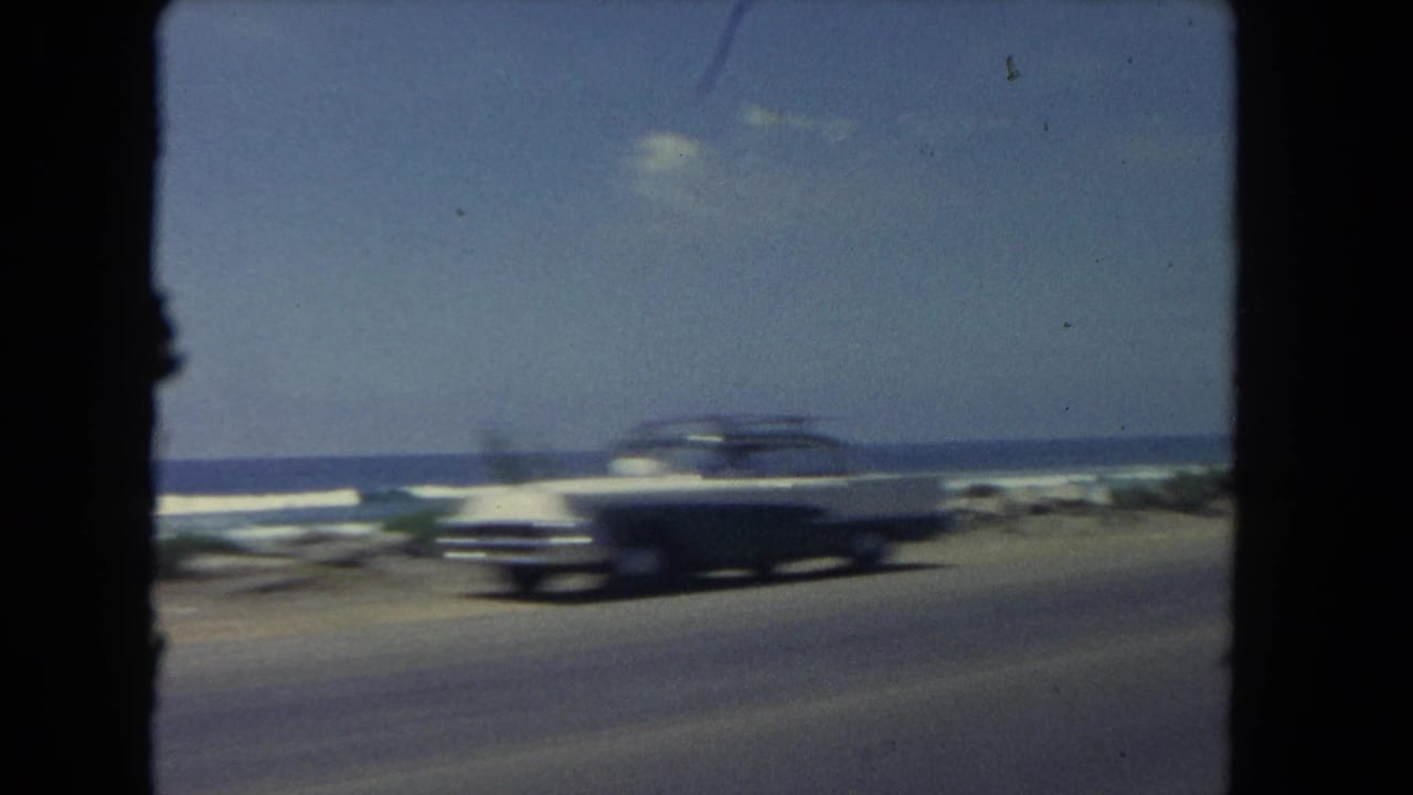Classic dark sedan passes on a coastal road with ocean and waves behind it.
