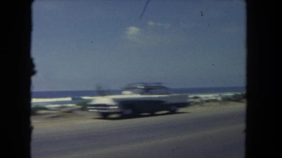 A dark mid-century sedan moves past the camera on a Los Angeles coastal road, with surf and a bright horizon behind it.