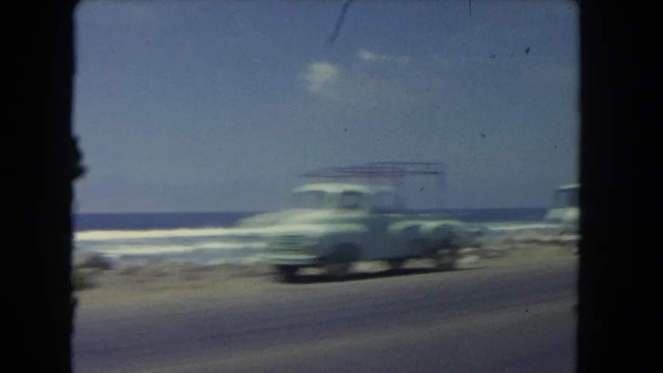 A light-colored van or wagon blurs past on a 1962 Los Angeles coastal road, with waves and shoreline visible in the background.