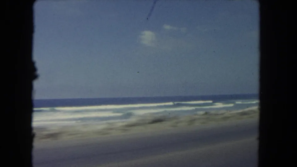 The moving-car view opens briefly to the Los Angeles shoreline, showing ocean waves, a low roadside edge, and blue sky without visible people.