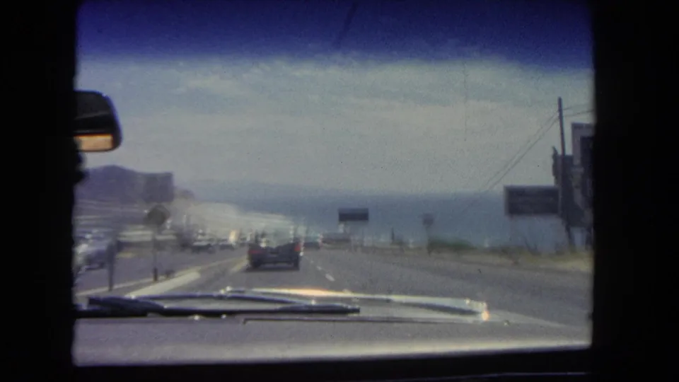A 1962 passenger-view frame shows a car following traffic on a Los Angeles coastal highway with ocean water and roadside signs in view.