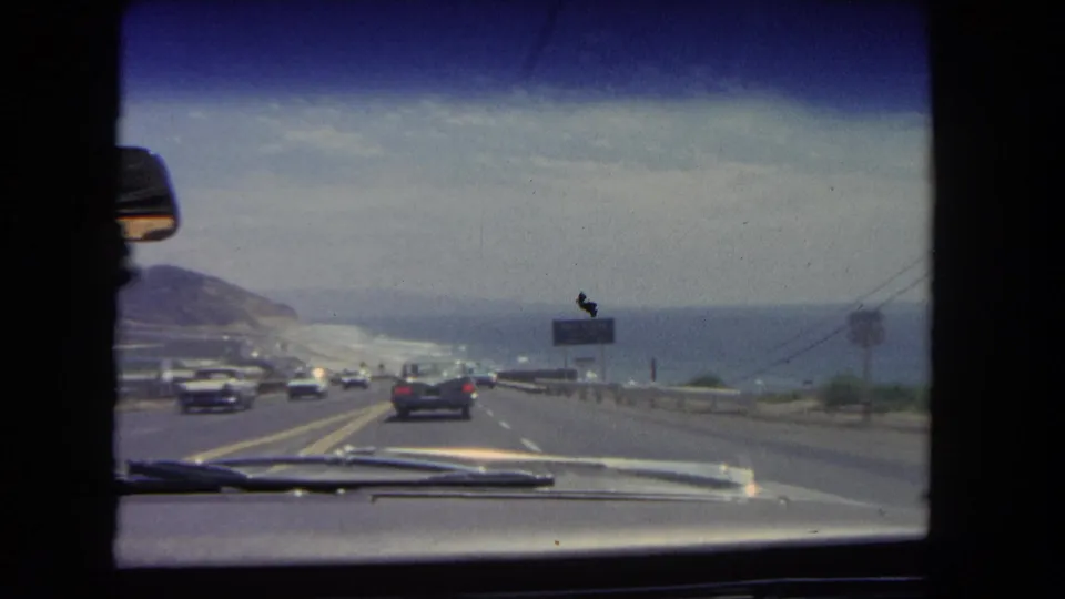 Passenger-view 8mm footage from 1962 Los Angeles shows traffic descending along a coastal road with the ocean and roadside signage nearby.