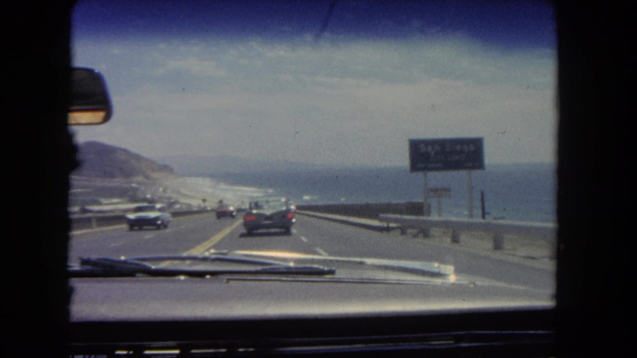 Coastal Los Angeles highway view from inside a moving car, with period automobiles ahead, a roadside sign, guardrail, ocean, and hills.