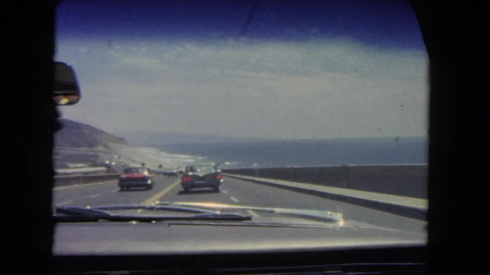 Vintage 8mm view of a 1962 coastal highway drive in Los Angeles, showing cars, lane markings, ocean water, hills, and the car windshield.
