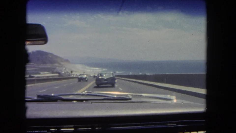 Interior windshield perspective of a 1962 Los Angeles coastal highway, with classic cars ahead, a guardrail, ocean to the right, and hills in the distance.