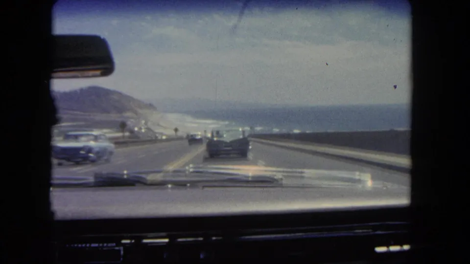 Windshield perspective of 1960s traffic on a Los Angeles coastal highway, with hills to the left and ocean to the right.