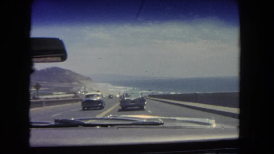 Interior car view from 1962 footage showing highway traffic, coastal hills, and the Pacific horizon near Los Angeles.