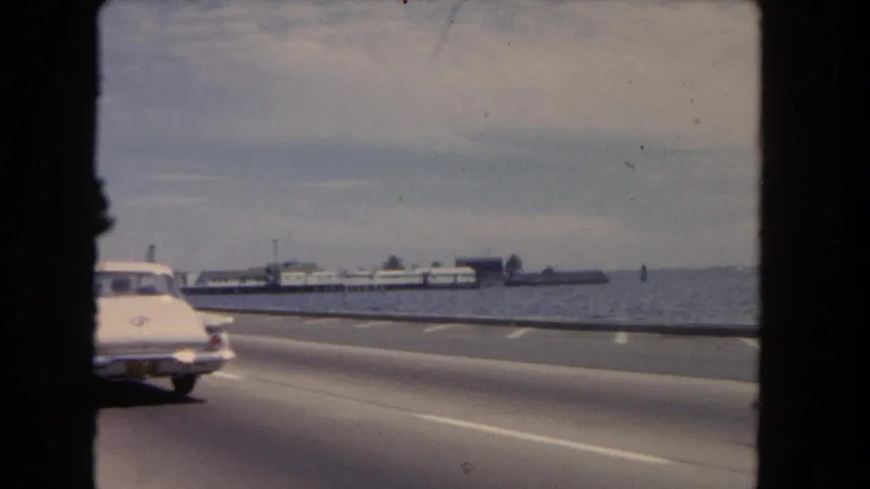 1962 8mm coastal road view in Los Angeles metadata, showing an older car, water, roadway, and a distant pier-like structure.