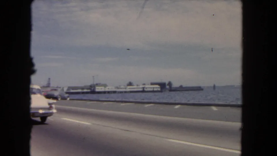Passenger-side 8mm view of a coastal Los Angeles road, with mid-century cars, open water, and a distant pier-like form.