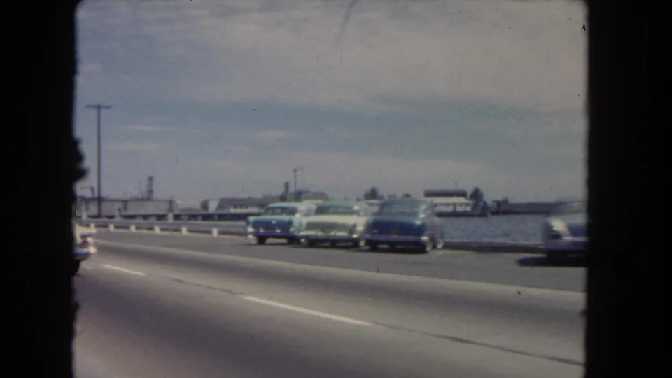 1962 home-movie view of cars traveling along a waterfront road in Los Angeles, seen through the edge of the film frame.
